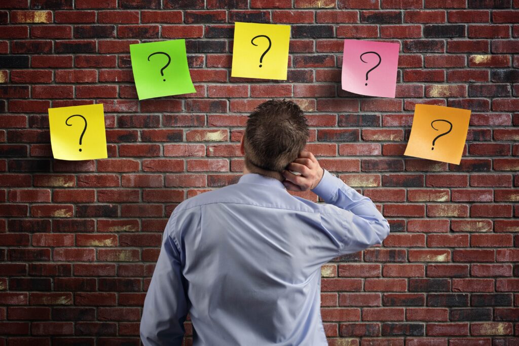 Man scratching his head while looking at colorful sticky notes with question marks on a brick wall